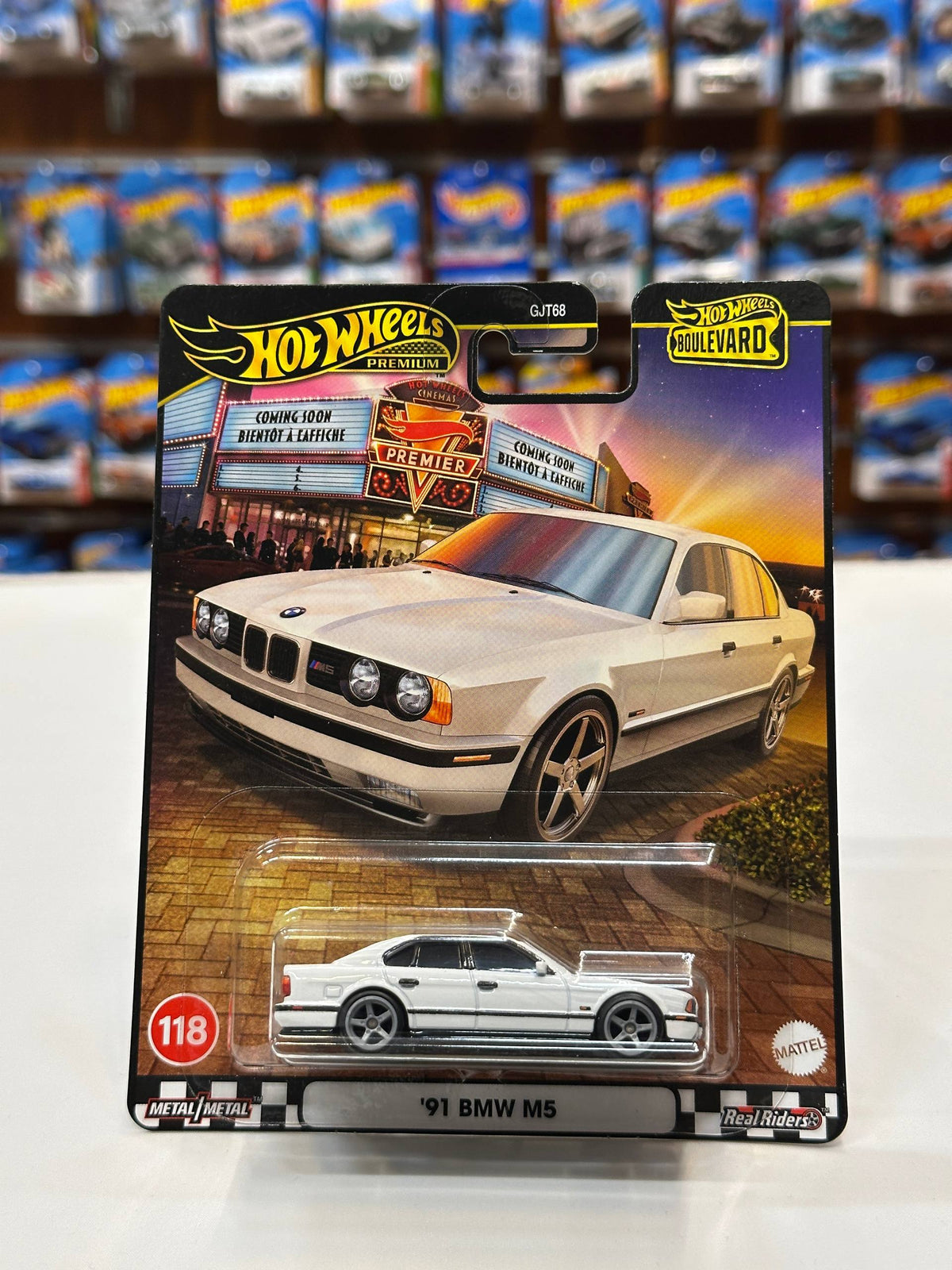 A Hot Wheels die-cast model car of a '91 BMW M5, packaged on its original card. The card features a graphic of a cream-colored BMW M5 parked in front of the 'Biker Shop' from the movie Hot Wheels.