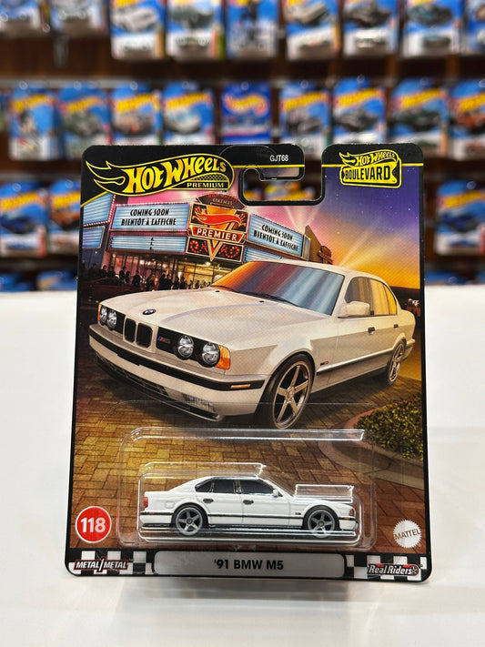 A Hot Wheels die-cast model car of a '91 BMW M5, packaged on its original card. The card features a graphic of a cream-colored BMW M5 parked in front of the 'Biker Shop' from the movie Hot Wheels.